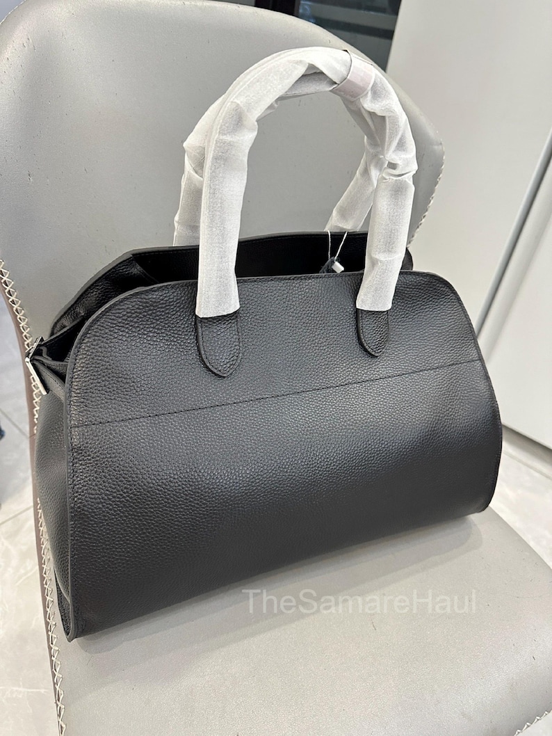 May include: A black leather handbag with a textured finish. The bag has a rounded shape, a top handle wrapped in white paper, and a silver chain detail on the side. The bag is sitting on a gray chair.