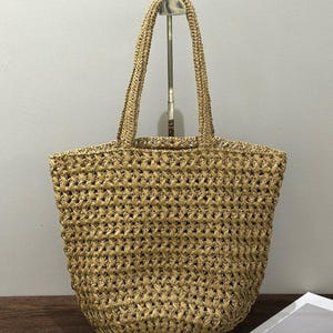May include: A woven straw tote bag with two handles. The bag is a light tan colour and has a textured, open weave design. The handles are made of the same material as the bag. The bag is empty and sitting upright.