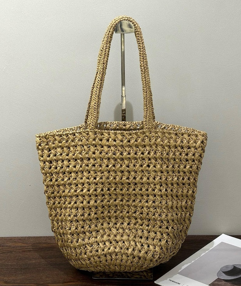 May include: A woven straw tote bag with two handles. The bag is a light tan colour and has a textured, open weave design. The handles are made of the same material as the bag. The bag is empty and sitting upright.