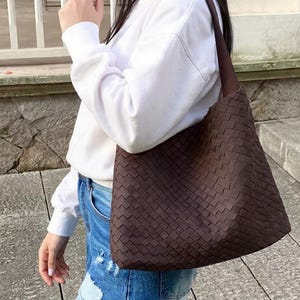 May include: A brown woven tote bag with a shoulder strap. The bag has a rectangular shape and a textured, basket-weave design. The bag is carried over the shoulder.