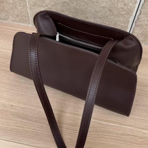 May include: A dark brown rectangular handbag with a long strap. The bag is open, revealing a zippered compartment. The stitching is visible along the edges of the bag and strap. The bag is sitting on a wooden surface.