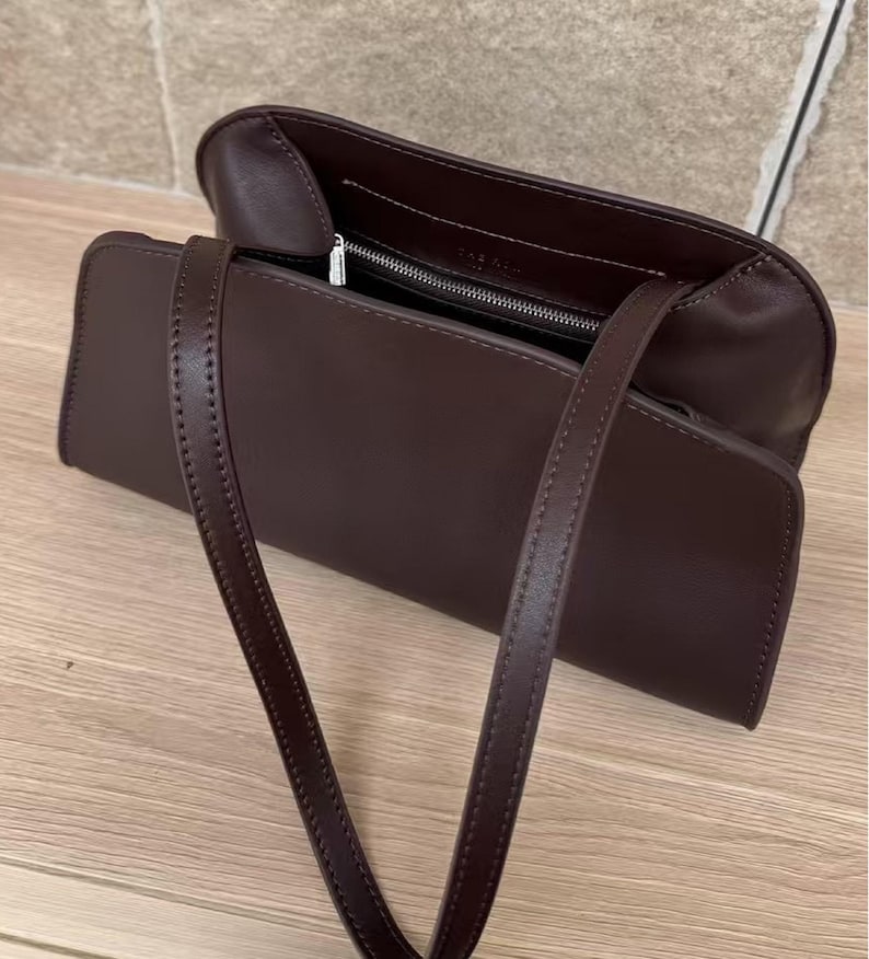 May include: A dark brown rectangular handbag with a long strap. The bag is open, revealing a zippered compartment. The stitching is visible along the edges of the bag and strap. The bag is sitting on a wooden surface.