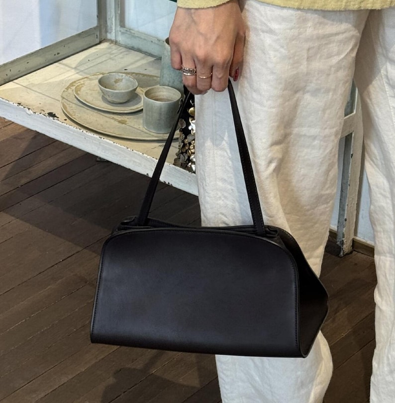 May include: A black rectangular handbag with a curved top and thin straps. The bag is held by a person wearing white trousers and a yellow top. The background includes a wooden floor and a shelf with ceramic items.