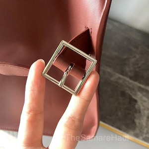 May include: Close-up of a burgundy-colored handbag with a silver square buckle. The bag's leather has a smooth texture. The buckle is attached to a strap that is part of the bag's design. The SamareHaul is written on the bottom right.