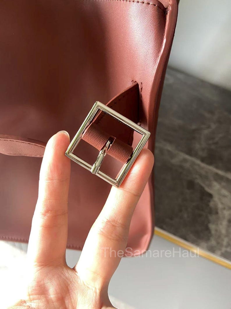 May include: Close-up of a burgundy-colored handbag with a silver square buckle. The bag's leather has a smooth texture. The buckle is attached to a strap that is part of the bag's design. The SamareHaul is written on the bottom right.