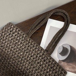 May include: A brown woven tote bag with braided handles. The bag features a basket-weave texture. The handles are a darker shade of brown. The bag is on a wooden surface next to a printed image of a hat.