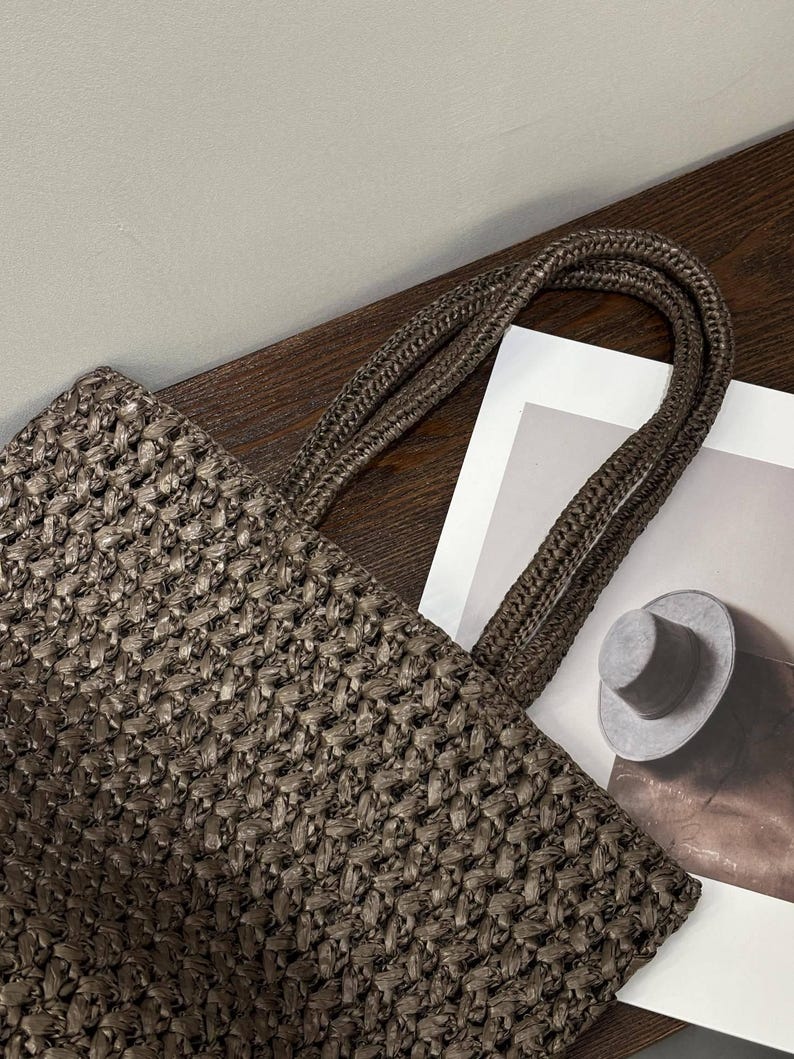 May include: A brown woven tote bag with braided handles. The bag features a basket-weave texture. The handles are a darker shade of brown. The bag is on a wooden surface next to a printed image of a hat.