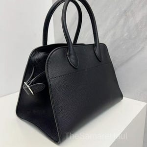 May include: A black handbag with two rounded handles. The bag has a rectangular shape with a textured surface and a silver-colored clasp. The bag is sitting on a white surface.