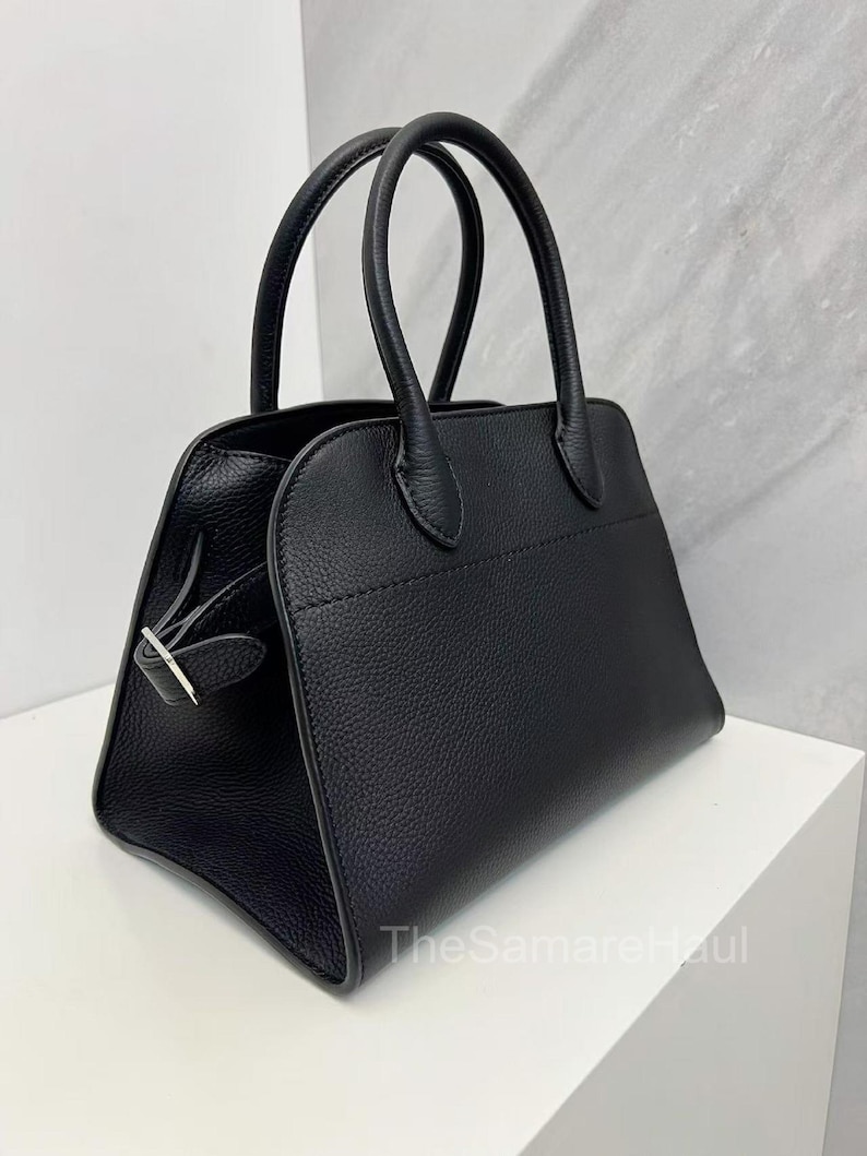 May include: A black handbag with two rounded handles. The bag has a rectangular shape with a textured surface and a silver-colored clasp. The bag is sitting on a white surface.