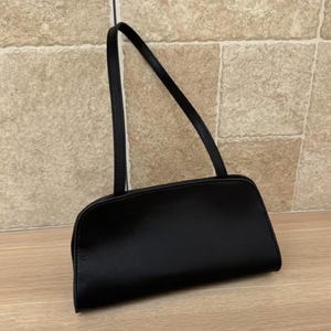 May include: A black rectangular handbag with a long, thin strap. The bag has a rounded bottom and a smooth, shiny surface. The strap is also black and appears to be made of the same material as the bag. The bag is sitting on a wooden surface.