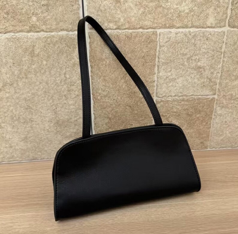 May include: A black rectangular handbag with a long, thin strap. The bag has a rounded bottom and a smooth, shiny surface. The strap is also black and appears to be made of the same material as the bag. The bag is sitting on a wooden surface.