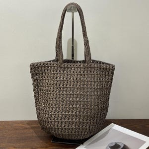 May include: A brown woven tote bag with two handles. The bag has a basket-like weave and a rectangular shape. The handles are also woven. A magazine with a picture of a hat is visible in the foreground. The bag is suitable for carrying items.