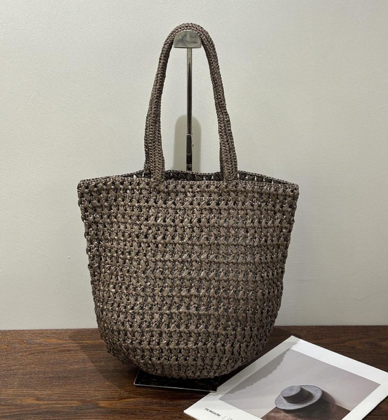 May include: A brown woven tote bag with two handles. The bag has a basket-like weave and a rectangular shape. The handles are also woven. A magazine with a picture of a hat is visible in the foreground. The bag is suitable for carrying items.