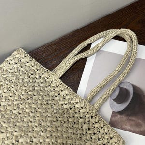 May include: A beige woven tote bag with matching rope handles. The bag has a textured, basket-weave pattern. The handles are thick and appear sturdy. The bag is partially visible, suggesting a larger size.