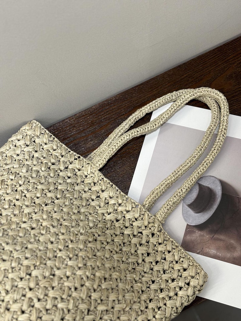 May include: A beige woven tote bag with matching rope handles. The bag has a textured, basket-weave pattern. The handles are thick and appear sturdy. The bag is partially visible, suggesting a larger size.