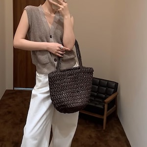 May include: A dark brown woven tote bag with two handles. The bag has a cylindrical shape and appears to be made of a natural fibre. The bag is being held by a person wearing a sleeveless, button-down vest and white trousers.