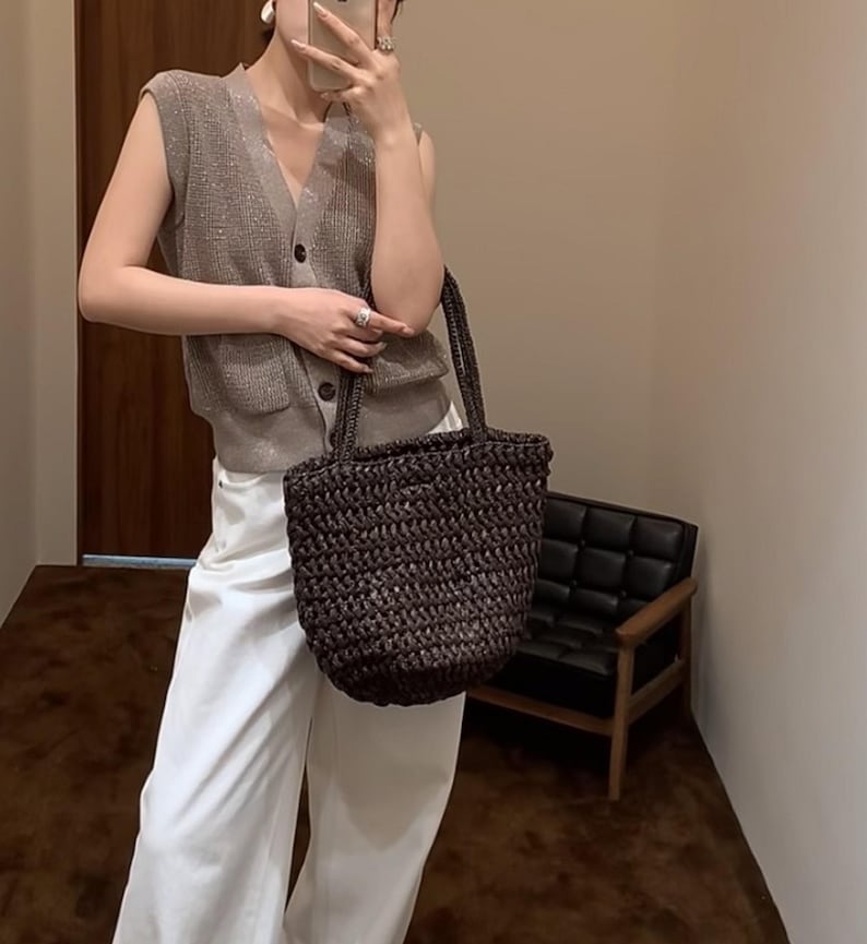May include: A dark brown woven tote bag with two handles. The bag has a cylindrical shape and appears to be made of a natural fibre. The bag is being held by a person wearing a sleeveless, button-down vest and white trousers.