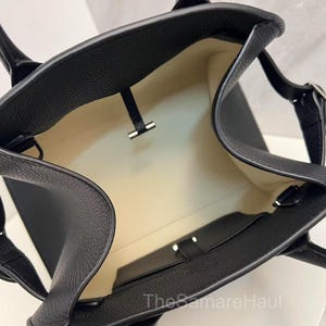 May include: Black leather tote bag with a textured finish. The bag has a light-colored interior and a small pocket. Features two handles and a silver-colored clasp. The bag is open, showing the inside.