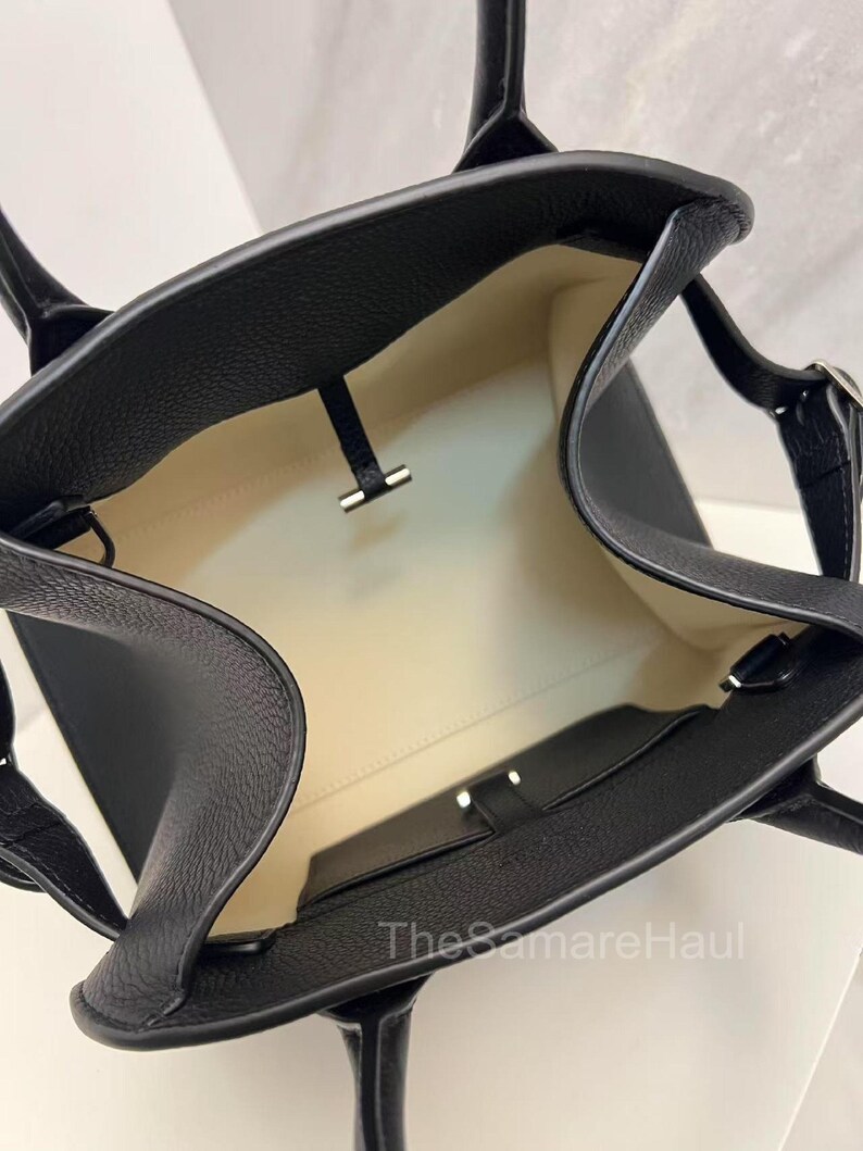 May include: Black leather tote bag with a textured finish. The bag has a light-colored interior and a small pocket. Features two handles and a silver-colored clasp. The bag is open, showing the inside.