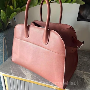May include: A dusty rose-colored handbag with two rounded handles and a rectangular shape. The bag has a flap closure with a silver-colored buckle. The bag is sitting on a gray and gold-trimmed surface. The text "TheSameHaul" is visible.