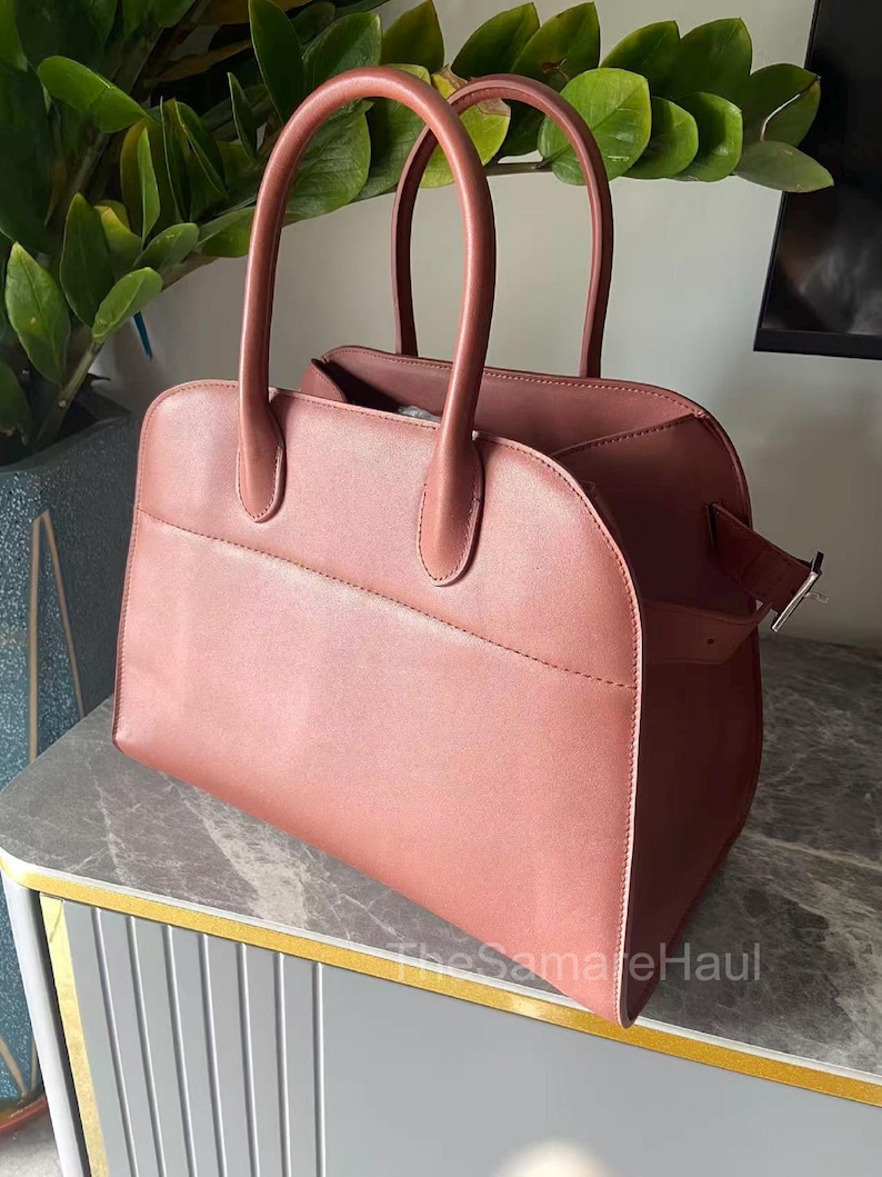 May include: A dusty rose-colored handbag with two rounded handles and a rectangular shape. The bag has a flap closure with a silver-colored buckle. The bag is sitting on a gray and gold-trimmed surface. The text "TheSameHaul" is visible.