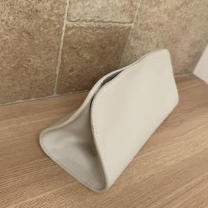 May include: A light gray, curved clutch bag with a minimalist design. The bag is made of a smooth material and features a subtle, stitched seam detail. The interior is a darker gray. The bag is sitting on a wooden surface.