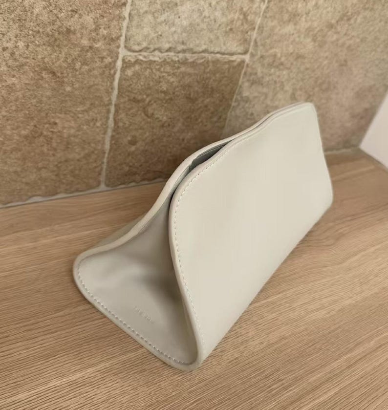 May include: A light gray, curved clutch bag with a minimalist design. The bag is made of a smooth material and features a subtle, stitched seam detail. The interior is a darker gray. The bag is sitting on a wooden surface.