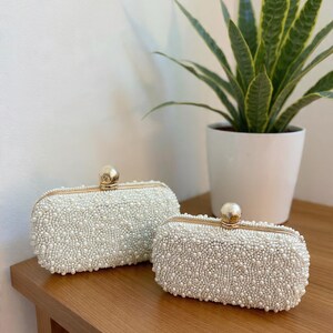 May include: Two rectangular, white beaded clutch bags with gold-toned frames and closures. The bags are displayed on a wooden surface, with a potted plant in the background. The purses are covered in small, round beads.