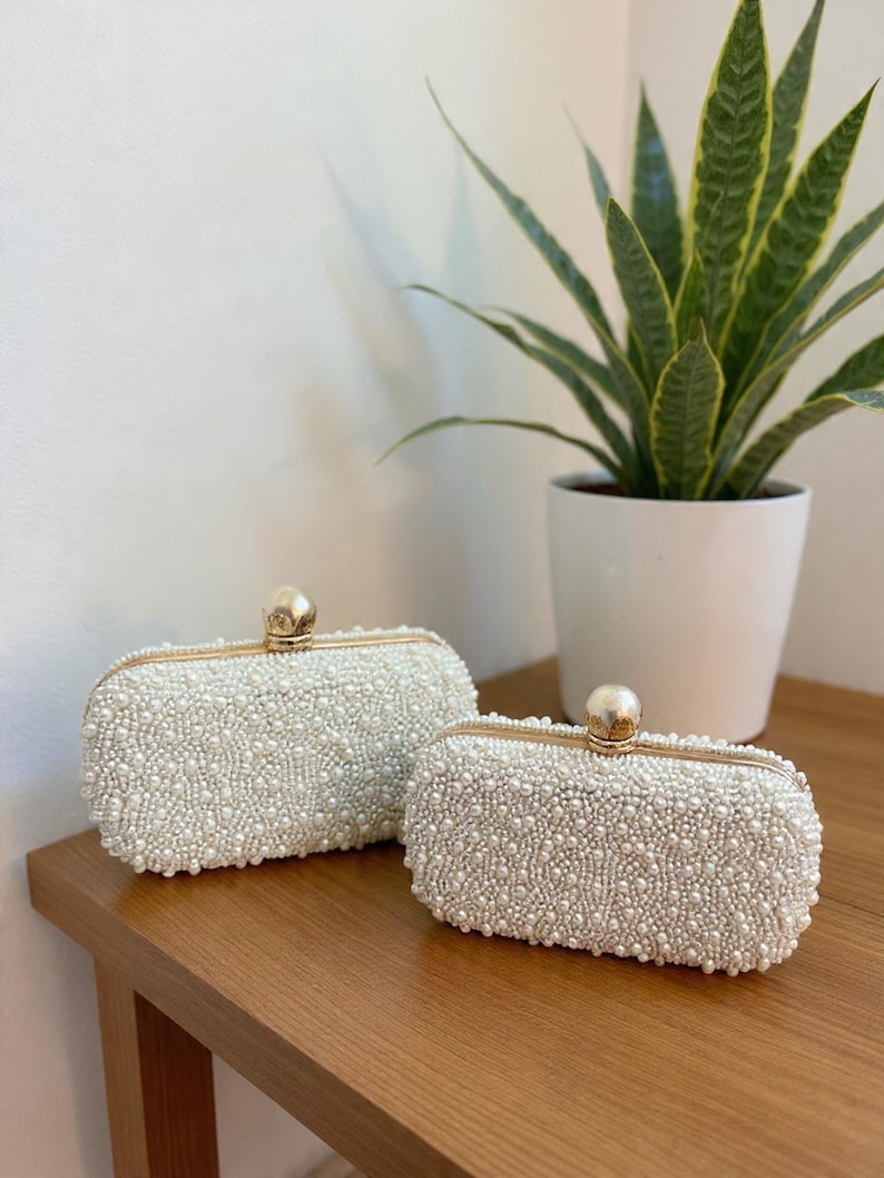 May include: Two rectangular, white beaded clutch bags with gold-toned frames and closures. The bags are displayed on a wooden surface, with a potted plant in the background. The purses are covered in small, round beads.