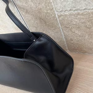 May include: Black leather handbag with a long, thin strap. The bag has a curved, trapezoidal shape and a minimalist design. The interior is visible, revealing a pocket. The bag is set against a neutral background.