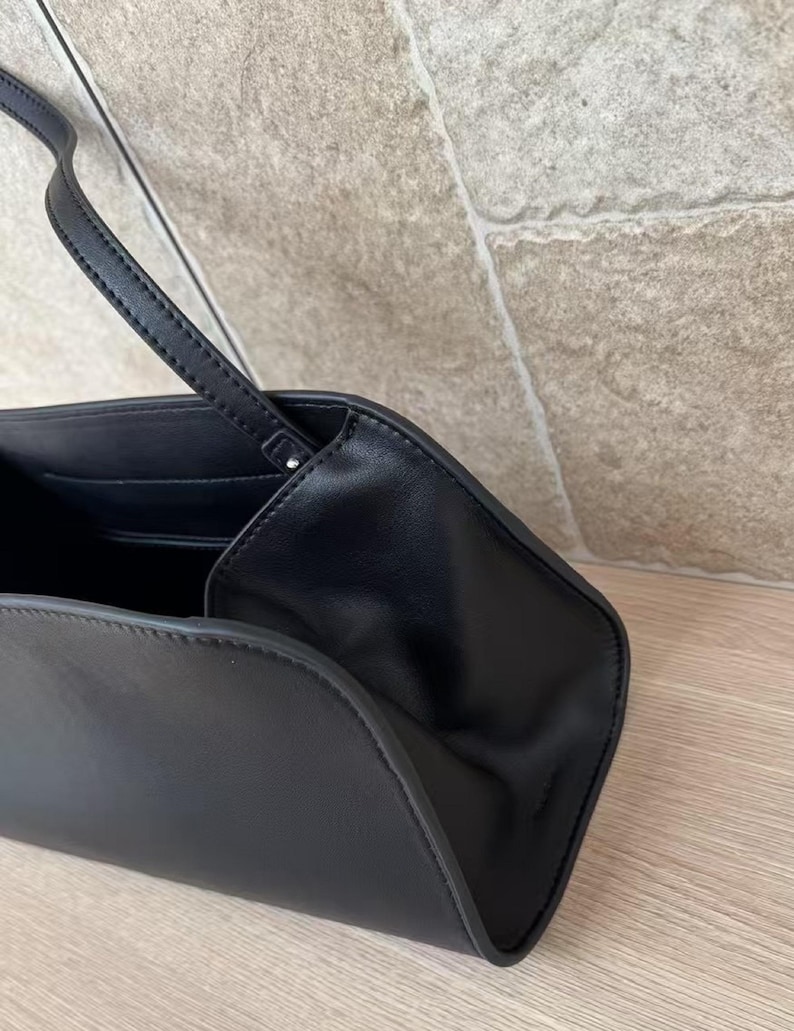 May include: Black leather handbag with a long, thin strap. The bag has a curved, trapezoidal shape and a minimalist design. The interior is visible, revealing a pocket. The bag is set against a neutral background.