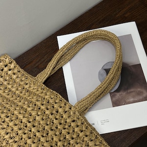 May include: A woven straw tote bag with sturdy handles. The bag has a textured, open weave design and is tan in colour. The handles are made of the same material and are tightly woven. The bag is on a dark brown surface.
