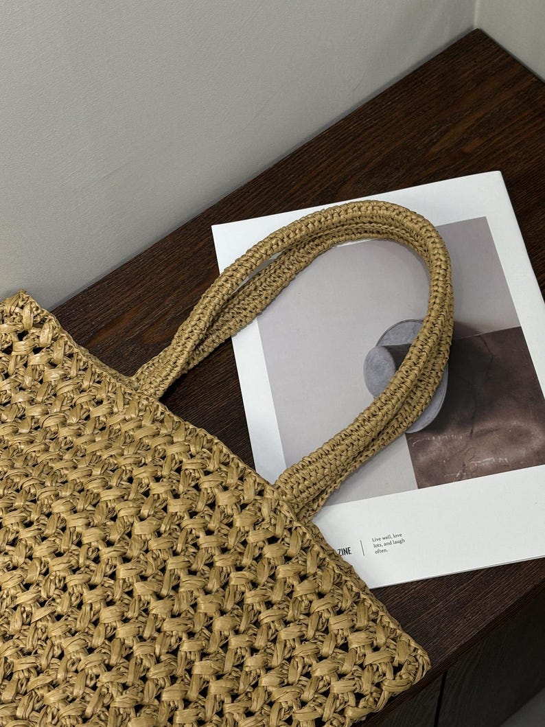 May include: A woven straw tote bag with sturdy handles. The bag has a textured, open weave design and is tan in colour. The handles are made of the same material and are tightly woven. The bag is on a dark brown surface.