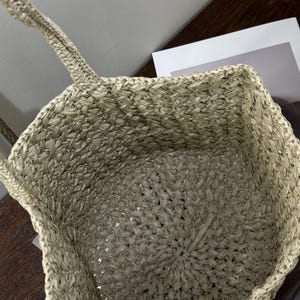 May include: A beige woven tote bag with two handles. The bag's interior is visible, showing a spacious design. The weave creates a textured surface. The bag is designed for carrying items and has a casual, everyday style.