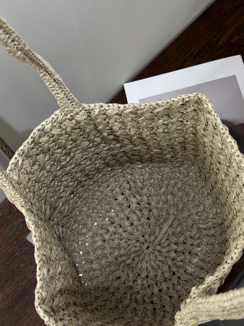 May include: A beige woven tote bag with two handles. The bag's interior is visible, showing a spacious design. The weave creates a textured surface. The bag is designed for carrying items and has a casual, everyday style.