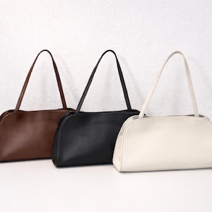 May include: Three shoulder bags in brown, black, and cream. Each bag has a rectangular shape with rounded corners and a long, thin strap. The bags are made of a smooth material and are displayed against a white background.