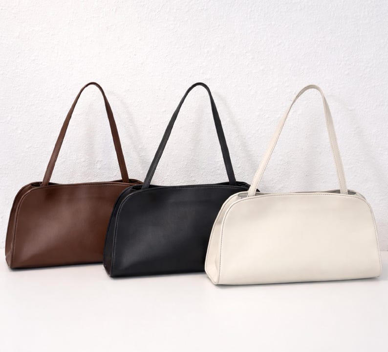 May include: Three shoulder bags in brown, black, and cream. Each bag has a rectangular shape with rounded corners and a long, thin strap. The bags are made of a smooth material and are displayed against a white background.