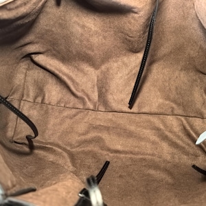 Puede incluir: Vista interior de un bolso tote negro con forro de tela marr&oacute;n. El bolso tiene correas negras y una etiqueta adjunta. El forro es de color marr&oacute;n liso y el bolso parece estar vac&iacute;o.