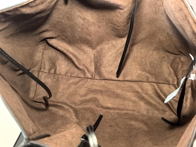 Puede incluir: Vista interior de un bolso tote negro con forro de tela marr&oacute;n. El bolso tiene correas negras y una etiqueta adjunta. El forro es de color marr&oacute;n liso y el bolso parece estar vac&iacute;o.
