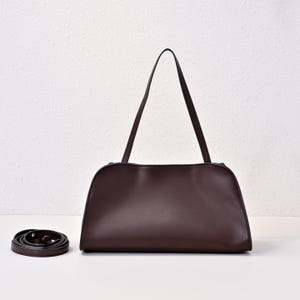 May include: A dark brown leather handbag with a rectangular shape and two long handles. A detachable shoulder strap is coiled on the left. The bag has a minimalist design and a smooth texture, suitable for everyday use.
