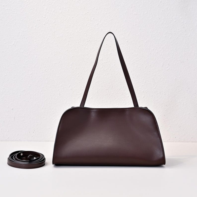 May include: A dark brown leather handbag with a rectangular shape and two long handles. A detachable shoulder strap is coiled on the left. The bag has a minimalist design and a smooth texture, suitable for everyday use.