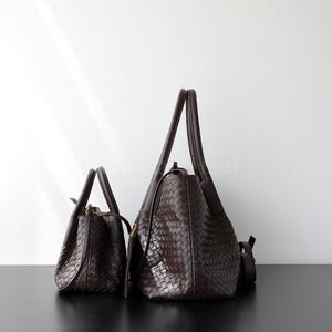 Puede incluir: Dos bolsos de cuero marr&oacute;n oscuro con textura tejida. Uno es un bolso tote m&aacute;s peque&ntilde;o con un asa superior, y el otro es un bolso de hombro m&aacute;s grande con una correa larga. Ambos bolsos est&aacute;n sobre una superficie negra.