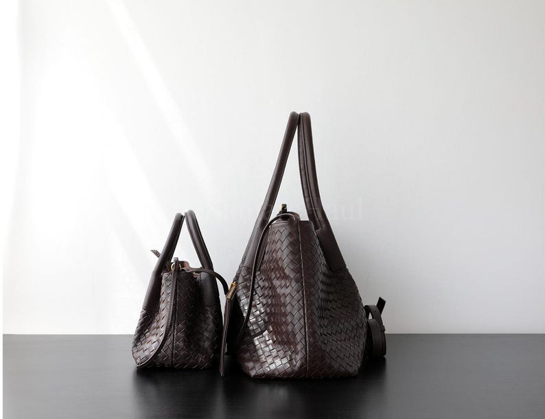 Puede incluir: Dos bolsos de cuero marr&oacute;n oscuro con textura tejida. Uno es un bolso tote m&aacute;s peque&ntilde;o con un asa superior, y el otro es un bolso de hombro m&aacute;s grande con una correa larga. Ambos bolsos est&aacute;n sobre una superficie negra.