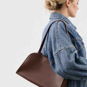 May include: A brown shoulder bag with a curved top and a rectangular base. The bag has a long strap and is made of a smooth material. The bag is being worn over a denim jacket.