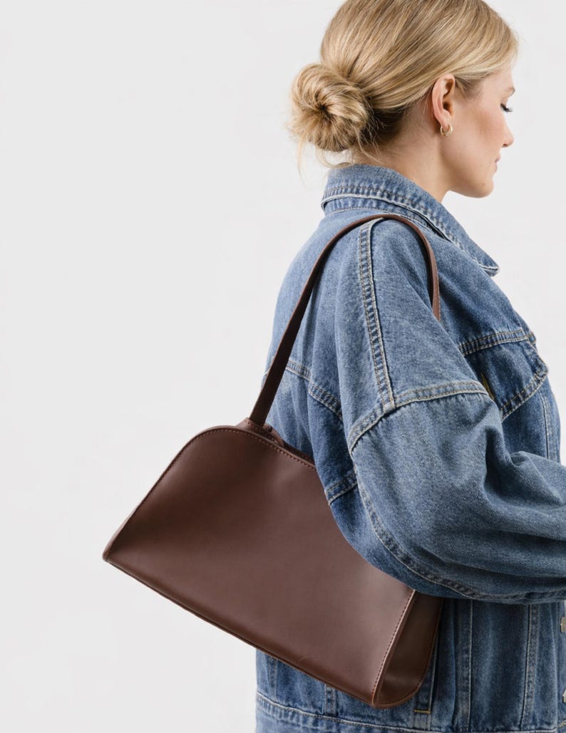 May include: A brown shoulder bag with a curved top and a rectangular base. The bag has a long strap and is made of a smooth material. The bag is being worn over a denim jacket.