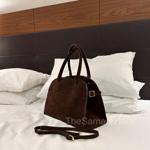 May include: A brown suede handbag with a top handle and a detachable shoulder strap. The bag has a rectangular shape with rounded edges and a gold-colored buckle closure. The bag is sitting on a white bed.