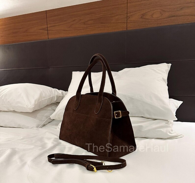 May include: A brown suede handbag with a top handle and a detachable shoulder strap. The bag has a rectangular shape with rounded edges and a gold-colored buckle closure. The bag is sitting on a white bed.