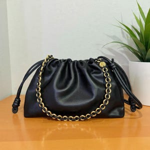 以下が含まれることがあります： 黒い革のハンドバッグ。ゴールドチェーンのストラップと巾着開閉式。バッグはギャザーデザインで、結び目のついた巾着が付いています。ゴールドチェーンはバッグの上部に巻き付けられています。バッグは木製の表面に置かれています。