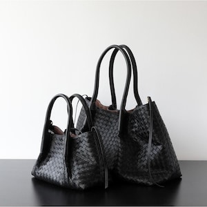 Puede incluir: Dos bolsos tote de cuero negro con textura tejida. Los bolsos tienen asas dobles y un dise&ntilde;o estructurado. Un bolso es m&aacute;s grande que el otro. Los bolsos est&aacute;n sobre una superficie negra contra un fondo blanco.