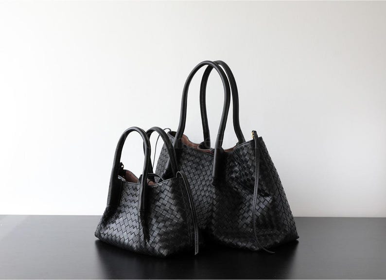 Puede incluir: Dos bolsos tote de cuero negro con textura tejida. Los bolsos tienen asas dobles y un dise&ntilde;o estructurado. Un bolso es m&aacute;s grande que el otro. Los bolsos est&aacute;n sobre una superficie negra contra un fondo blanco.